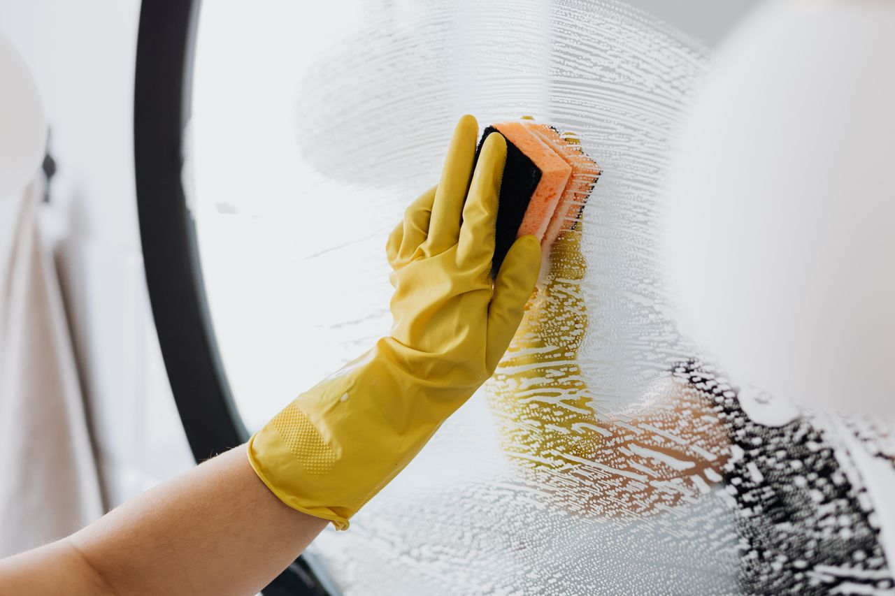 about-img Anonymous person in yellow rubber gloves using simple sponge for cleaning mirror in bathroom
