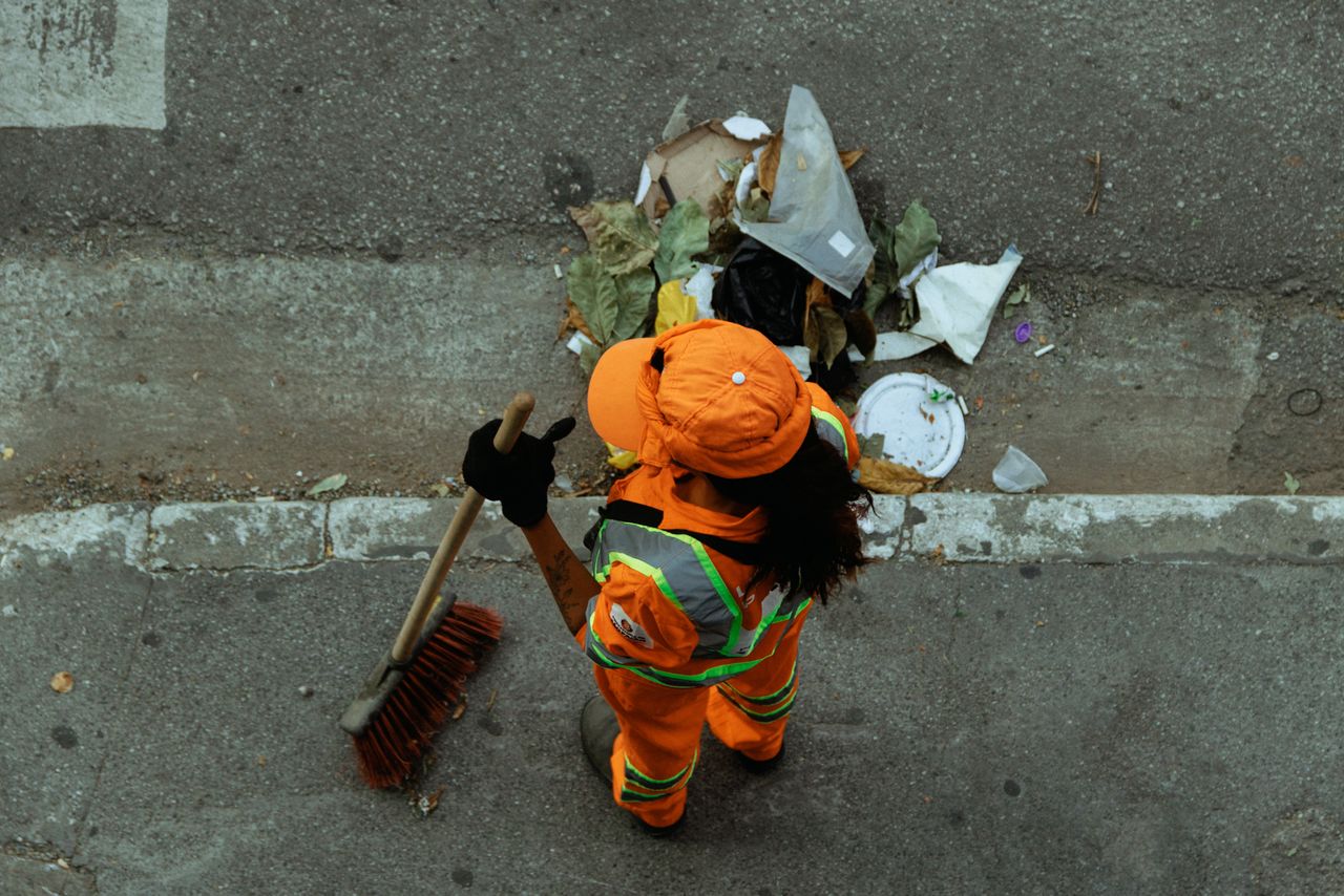 core-values Top view of a street cleaner wearing orange uniform sweeping debris on a city sidewalk.